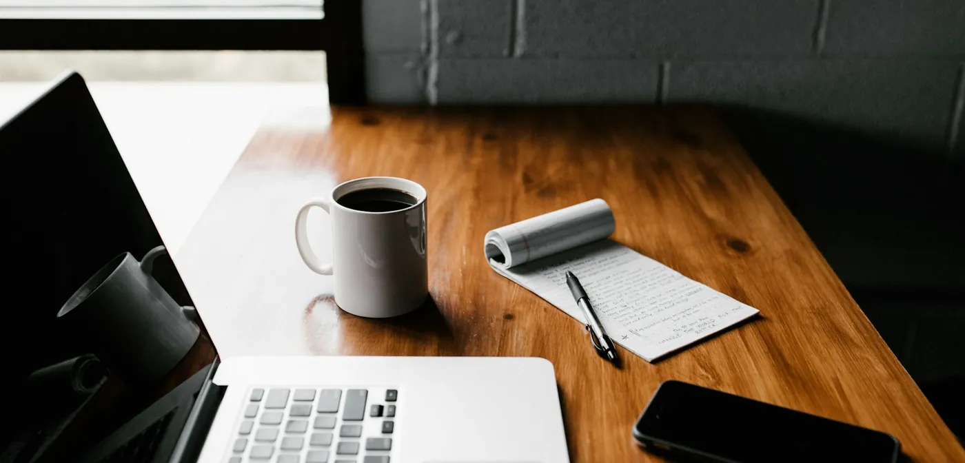 A wooden work table with a laptop, coffee mug, notepad, and pen.