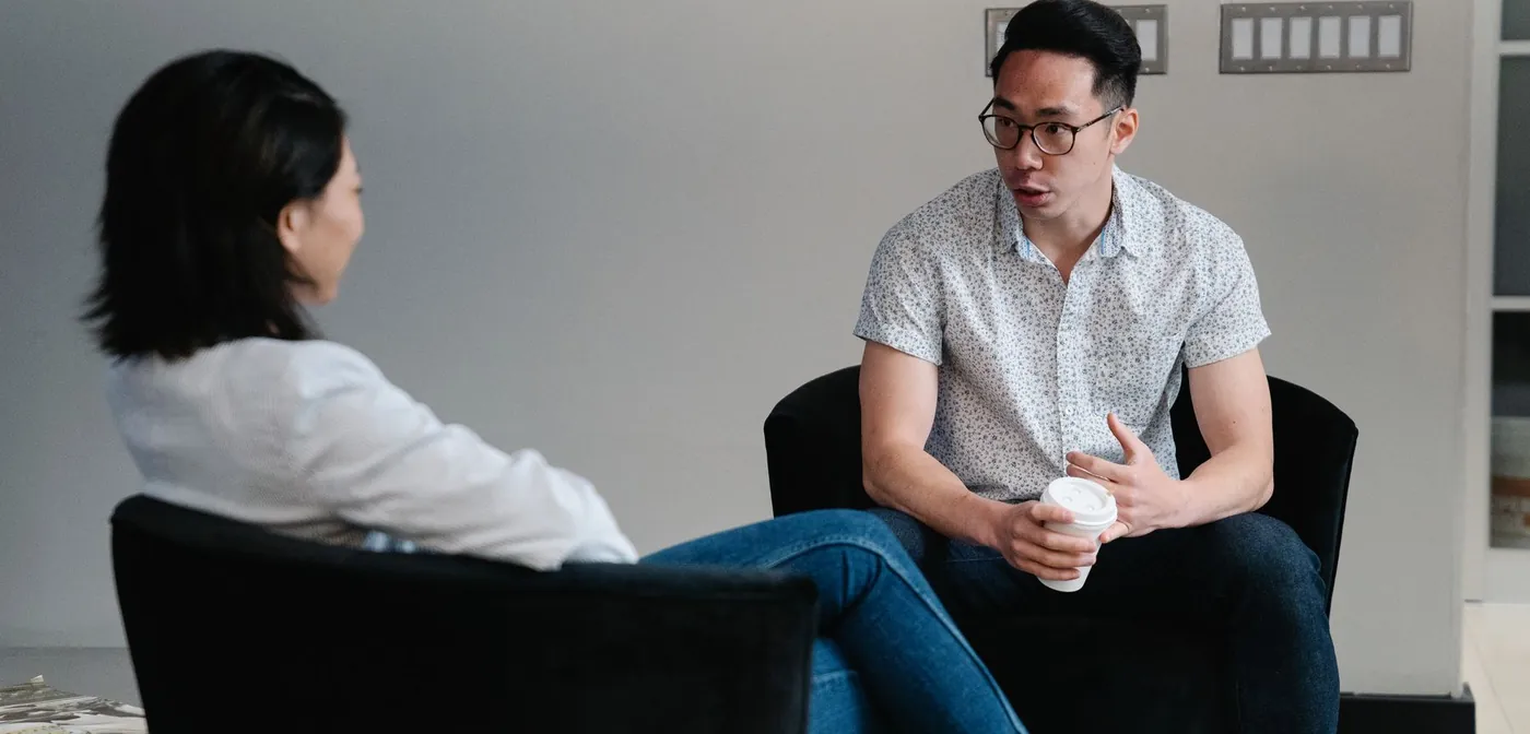 Two colleagues in a one-on-one conversation in lounge chairs, one listening while the other talks.